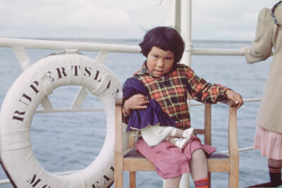 A young girl holding a doll on a boat. The life preserver next to her reads, "RUPERTSLAND"