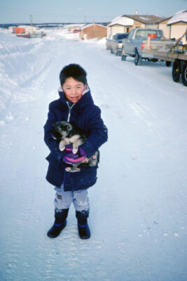 A young boy holding a little dog on a snowy road. Trucks and small buildings line the road.