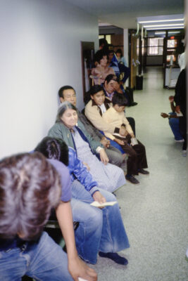 A group of people wait in a line in a hallway. Some people are sitting, some are standing.
