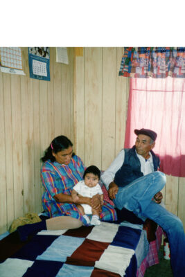 A man and woman holding a baby sit on a bed in a wood-panelled room. Three calendars hang on the wall above the woman.