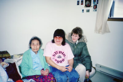 Three women sit on a bed. The woman in the middle wears a pink shirt that reads, "Studio 2000"