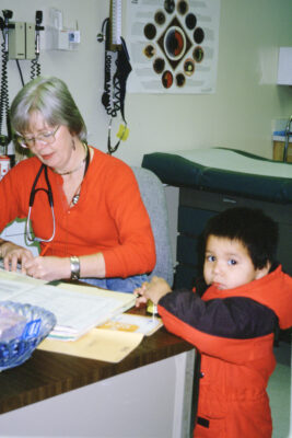 A woman in an orange shirt with a stethescope around her neck writes at a desk in a hospital examination room. A young boy in an orange and black jacket stands beside her and looks at the camera.