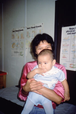 A woman in a pink shirt sits on a hospital examination table holding a baby.