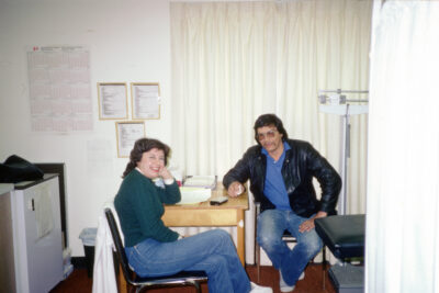 A man in a black leather jacket and a woman in a green sweater sit at a table in a hospital examination room.