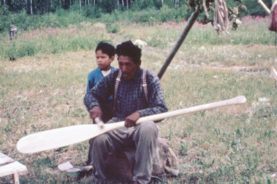 A man sits outside with an oar on his lap. A young boy stands behind him.