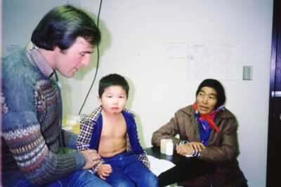 A boy with an injured chin sits on an examination table next to a man with a stethoscope. He has one man on the boy's arm and the other on the boy's back. A woman sits beside the table.
