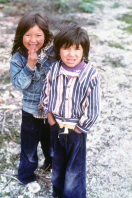 Two children stand outside, looking at the camera. One child wears a striped shirt and the other wears a jean jacket.