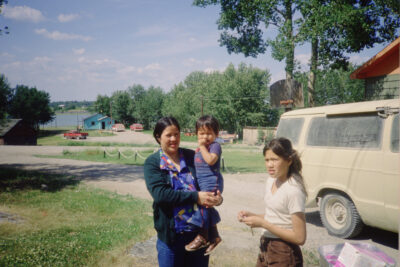 A woman holds a young boy and a girl stands beside them. They stand outdoors next to a beige van.