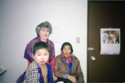 A boy with an injured chin sits on an examination table. A woman with a purple shawl stands behind him, and another woman sits beside her. A calendar hangs on the door next to them.