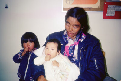 A woman in a dark blue jacket holds a baby in white clothing. A boy in a dark blue jacket stands beside them, sucking his thumb.