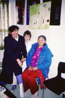 Three women sit in the corner of a room. One woman sits on another woman's lap. Posters hang on the wall above them.