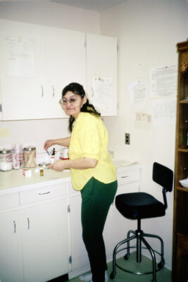 A woman in a yellow shirt stands at a counter with a pill bottle as she turns to the camera.
