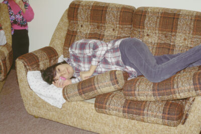A woman lies on a brown plaid couch holding an empty specimen container. A portion of a child eating an ice cream cone can be seen in the corner of the frame.