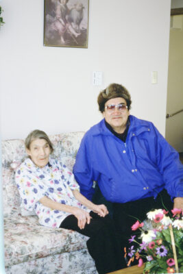 An elderly woman sits next to a man in a blue jacket and fur hat on a couch. Flowers sit on the table in front of them.