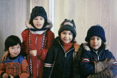 Four children standing together in front of a wood-panelled wall. Three children are in winter gear.