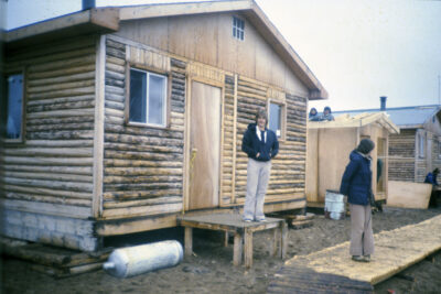 A man stands at the entrance of a log house. Another man stands on a log boardwalk, and two people can be seen in the background peering from the roof of a wooden structure that is being built.