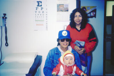 A man in a jean jacket, blue hat, and sunglasses holds a baby on his lap in a doctor's office. A woman in a red shirt stands behind him.