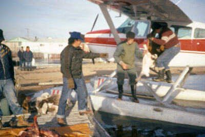 A man on a dock reaches for a person exiting a seaplane. Another person stands at the door of the seaplane. Other people can be seen watching in the distance.