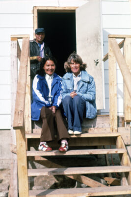 Two women sit on the steps of the entrance to a building. A man stands by the doorway behind them.