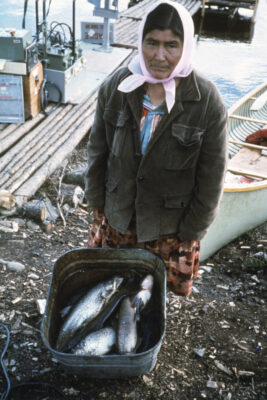 A woman with a scarf around her head stands next to a bucket of fish.