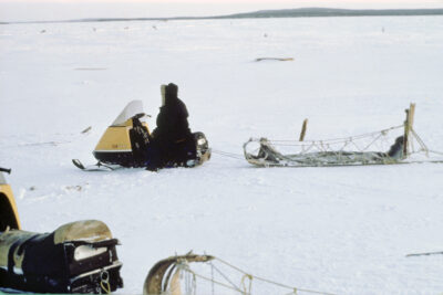 Two parked snowmobiles with sleds attached behind them on a snowy landscape. A person sits on one of the snowmobiles.