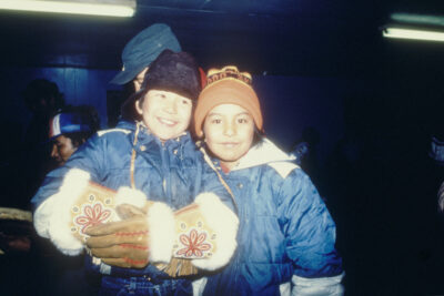 Two children in parkas and toques pose for the camera. One child wears beaded leather gauntlets.