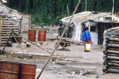 A woman holding a bucket walks amongst a cluster of log houses. Two dogs follow her. Red barrels are scattered throughout.