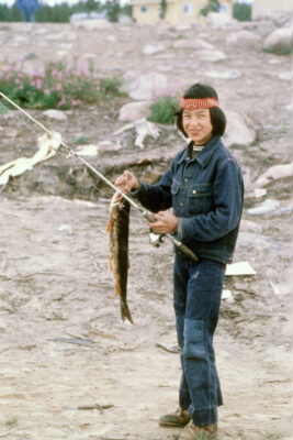 A boy wearing a jean outfit and a red headband holds a fish attached to a fishing pole.