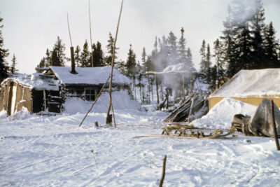 A snowy scene including a log house and a yellow tent. A sled sits next to the tent.
