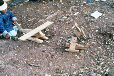 A child plays with a wooden airplane. Two other wooden airplanes sit on the ground nearby.
