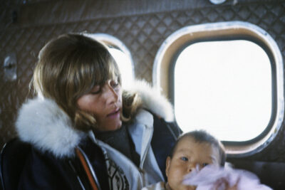 A woman in a coat with a white fur hood holds a baby on an airplane.