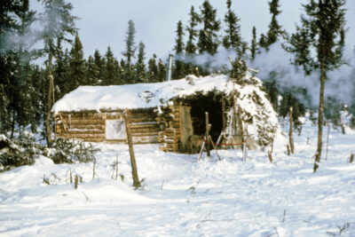 A snowy scene featuring a log house. Smoke billows from the chimney.