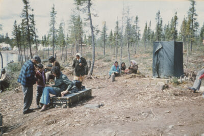 A group of people in a clearing of a forested area. Three women sit on the ground together in the background next to a tree, another person sits on a long black case next to a man and a boy, and a rectangular structure covered in a green tarp stands in the background.