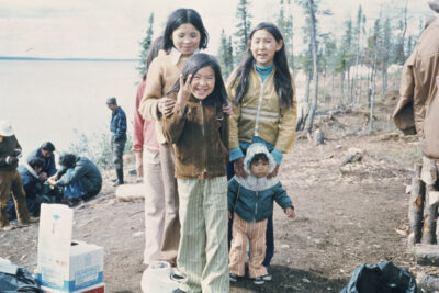 Four children and a toddler outdoors, gathered for the camera. A group of men can be seen crouched in the background.