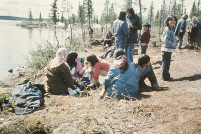 A group of people gathered by a lake. Some people sit together on the ground, others stand and chat with each other. One child stands with their hands in their jacket pockets and looks at the camera.