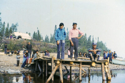 A group of people on the banks of a body of water. One man sits and two people stand at the end of a dock in the foreground. A white tent can be seen atop a small hill in the background.