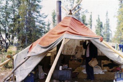 A tipi with an orange cover, with two people inside. A stove inside the tipi has a chimney that reaches out the top.