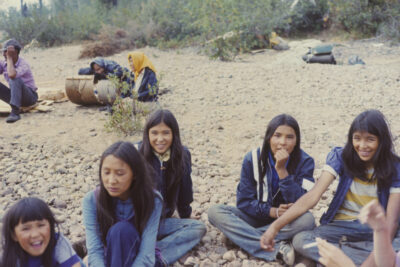 A group of children/teens sit on a pebbly ground in the foreground. Two women with scarves around their heads and a man in a purple shirt sit in the background.