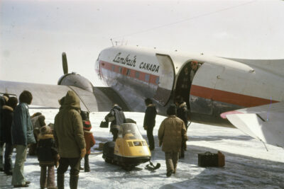 A group of people stand next to an airplane. A yellow snowmobile sits beside the plane, and two people can be seen in the doorway of the airplane.