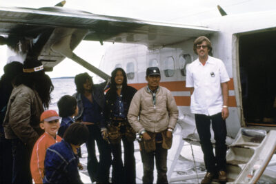 A group of people next to an airplane. A man in a white shirt stands on the stairs of the airpane.