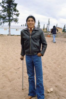 A man wearing a leather jacket, jeans, and a beaded neck piece leans on a cane on sandy ground.
