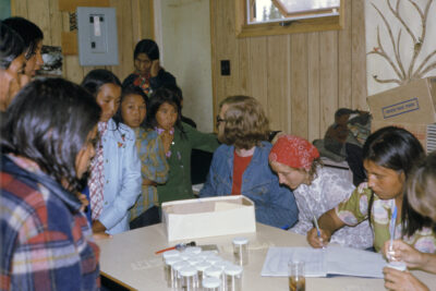 A group of people gathered around a table with empty specimen containers. A man in a jean jacket looks at a child in a green sweater. One woman writes in a book and another person writes on a specimen bottle. A woman stands in the corner of the room and looks at the camera.