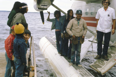 Three men stand on the float of a sea plane, one man in a white shirt stands on the airplane's stairs, and a group of people including three children stand on a dock beside the plane.