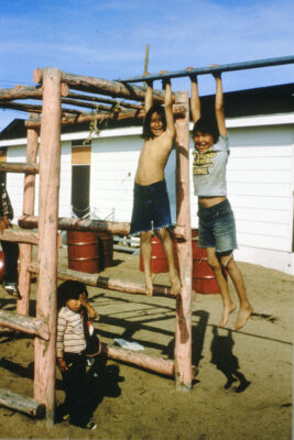 Two children hang from a bar attached to a wooden structure. A young child stands on the ground next to them with their hand on their left eye.