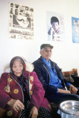 An elderly couple sits on a brown plaid couch. The woman wears a magenta embroidered coat, and the man has a cigarette in his mouth. Posters promoting health hang on the wall above them.