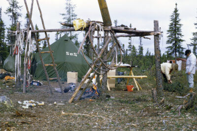 A structure made of tree branches stands in front of a green tent that reads "S.O.S." One person handles a rolled tarp while another person watches. A fire burns in the centre of the camp, and boxes, pots, and other camp materials scatter the site.