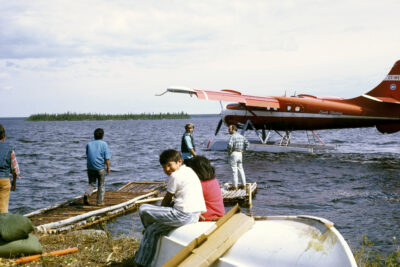 An orange sea plane floats on a body of water. Four men stand on a dock nearby, and two children sit on an upside-down boat on the shore.