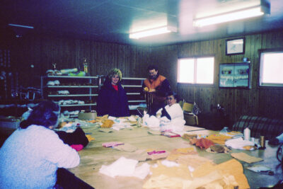 Five people around a large table covered with supplies for making moccasins. A shelf against the wall holds rows of finished moccasins.