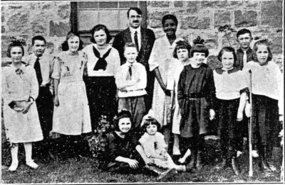 Ten children stand and two sit in front of a stone wall, with a man and a woman in the middle of the group.