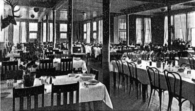 A room filled with multiple dining tables which are set with table cloths and dishes. A bust of an elk hangs on the wall on the left.